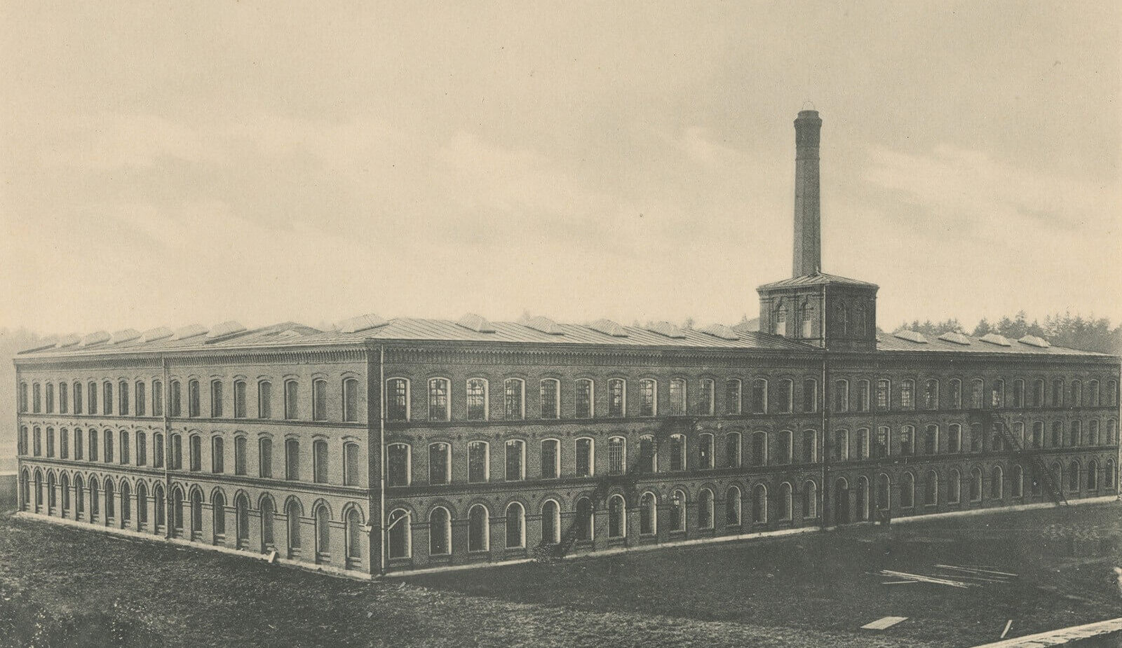 Zewnętrzny widok ceglanego budynku fabrycznego z wysokimi oknami i detalami z epoki