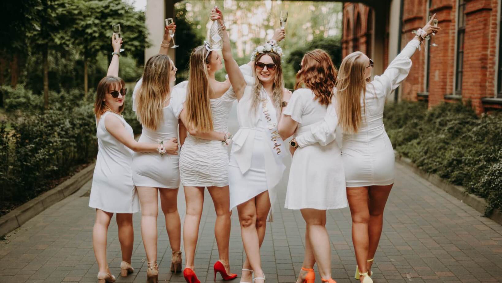 Group of friends dressed in white walking on the sidewalk celebrating the bride’s health with champagne