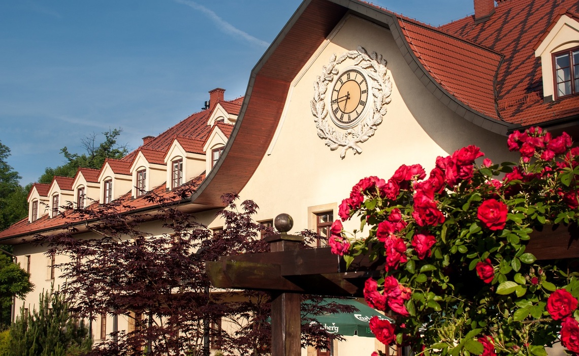 Historická fasáda hotelu s červenou taškovou střechou, vikýři a ozdobnými hodinami, v popředí červené růže a bujná zeleň.