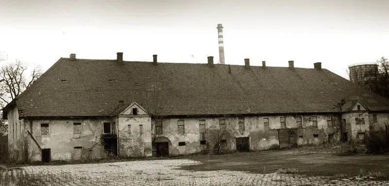 Dlouhá zchátralá stodola s taškovou střechou a malými okny, prázdné nádvoří a tovární komín v pozadí