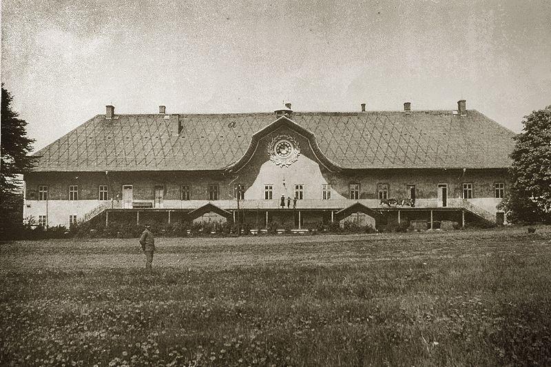 Historické sídlo s ozdobným středním štítem a kruhovým oknem, krytou terasou a širokým trávníkem s postavou.