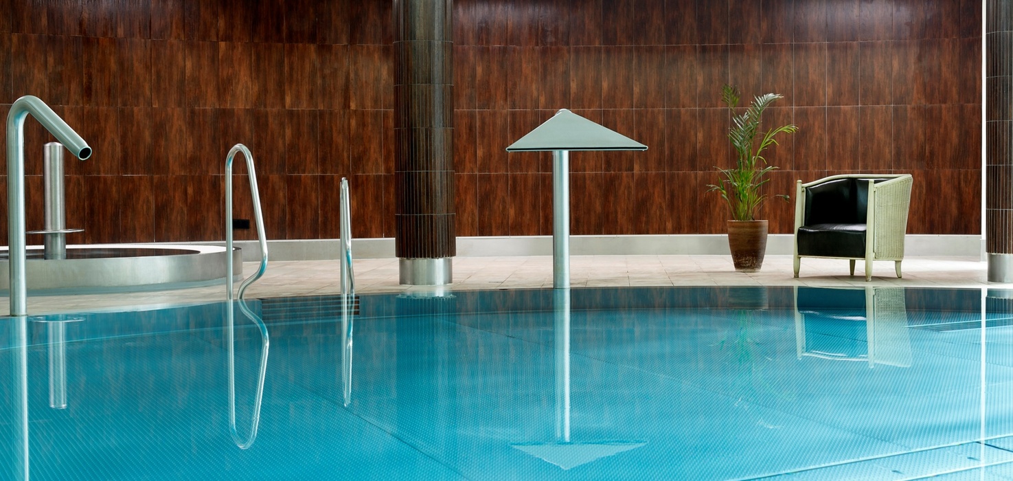 Vnitřní hotelový spa bazén se stříbrnými vodními tryskami, schůdky, proutěným křeslem a rostlinou před teplým dřevěným obložením