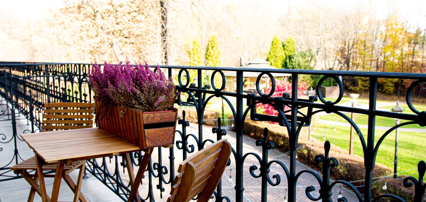 Kované zábradlí balkonu s dřevěným bistro stolem a židlemi, truhlík s fialovým vřesem, výhled na upravený park a altán.