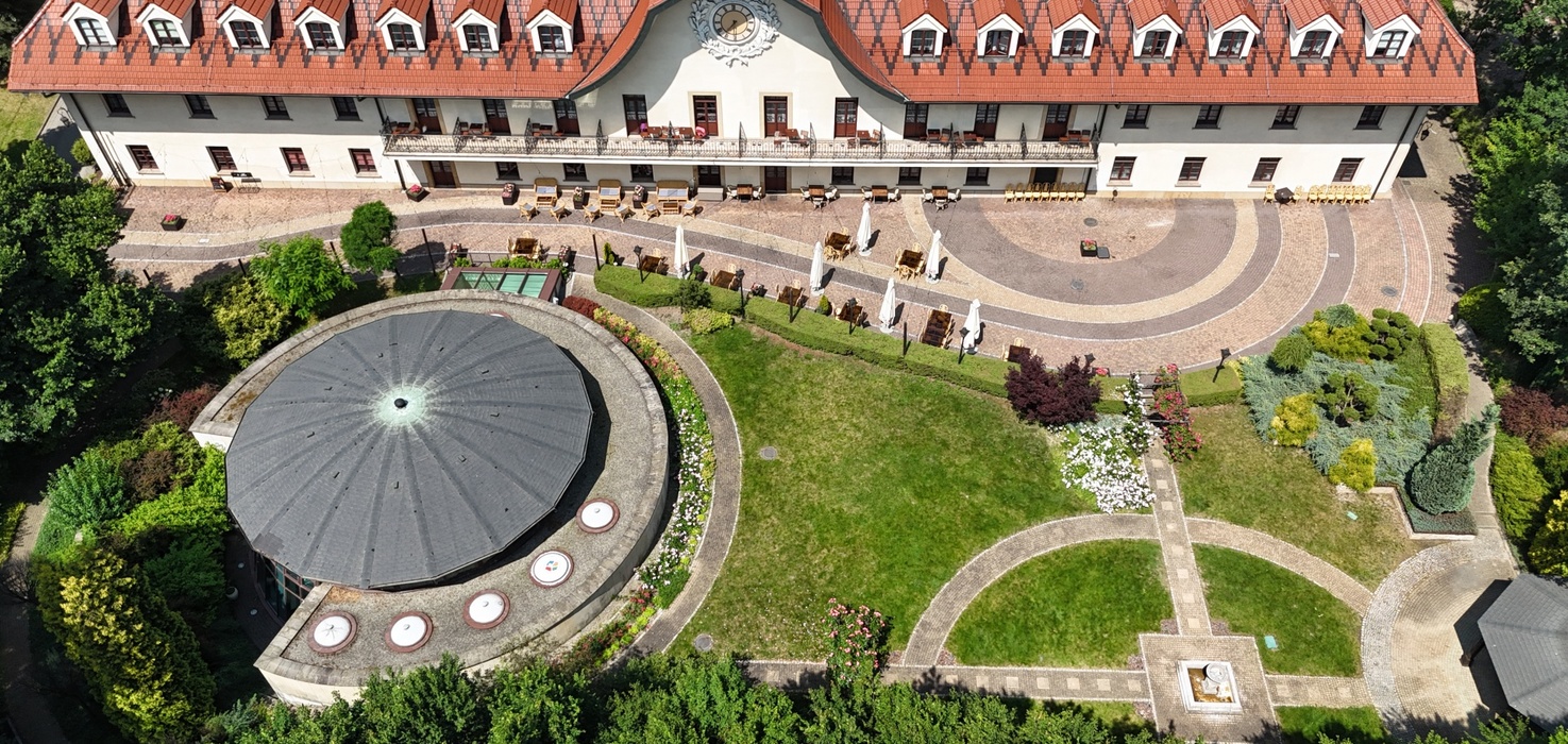 Historický hotel s červenou taškovou střechou a hodinami na fasádě, balkony a stolky na terase s výhledem na upravené kruhové zahrady a pavilon