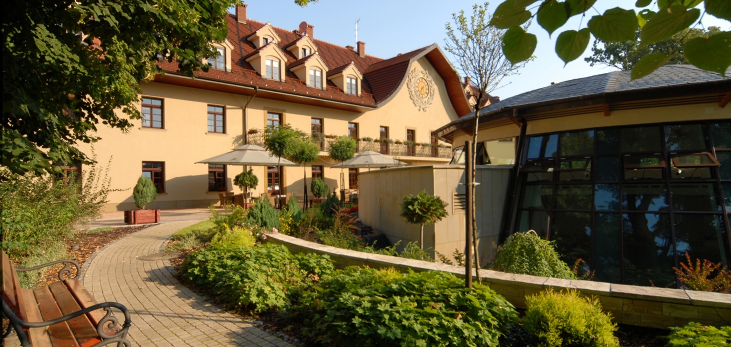 Malebné hotelové nádvoří se žlutou fasádou, červenou taškovou střechou, terasou s deštníky, posezením, upravenou zahradou a dlážděnou cestou.