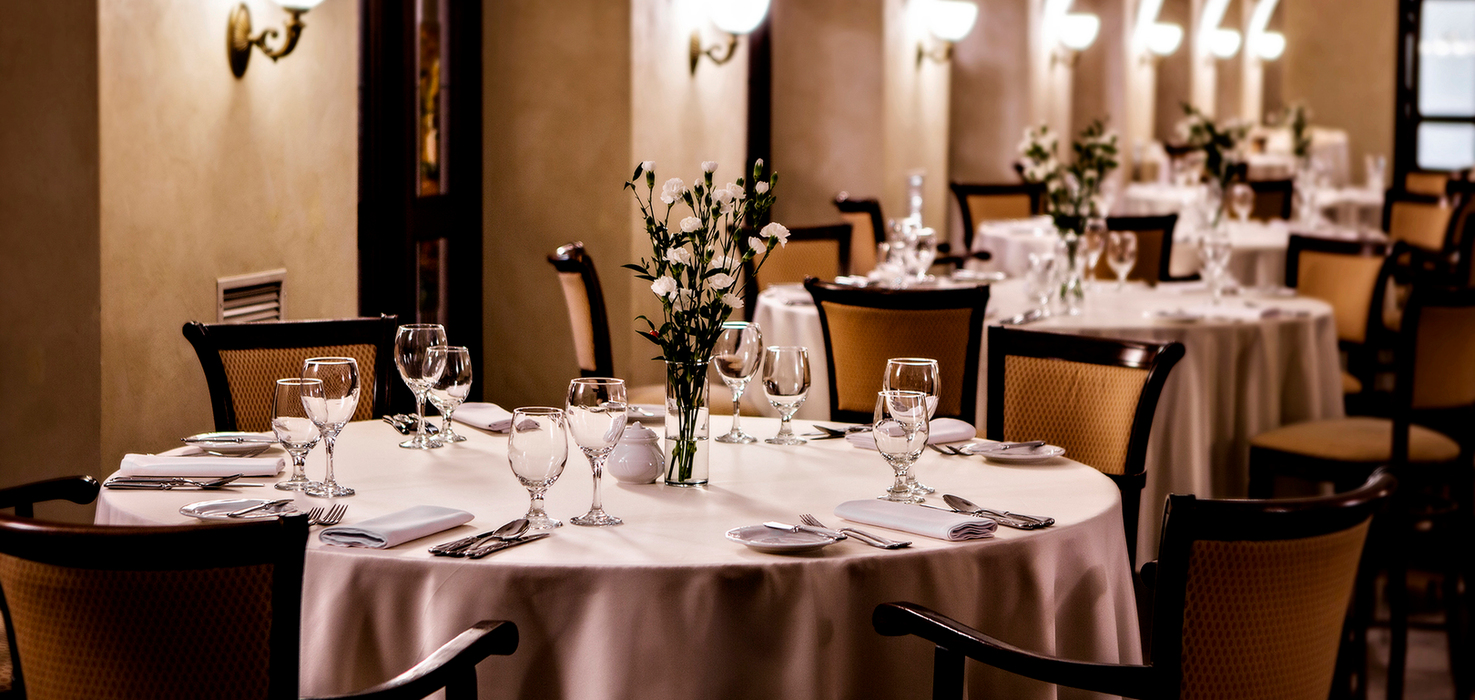Elegantní hotelová restaurace s kulatým stolem, bílým ubrusem, sklem a příbory, váza s bílými květy pod obloukovým, jemně osvětleným stropem