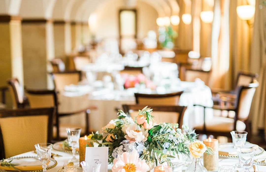 Elegantní banketová síň s obloukovými stropy, stůl s bílým ubrusem, květinovou dekorací, svíčkami, zlatým příborem a křišťálovými sklenicemi.