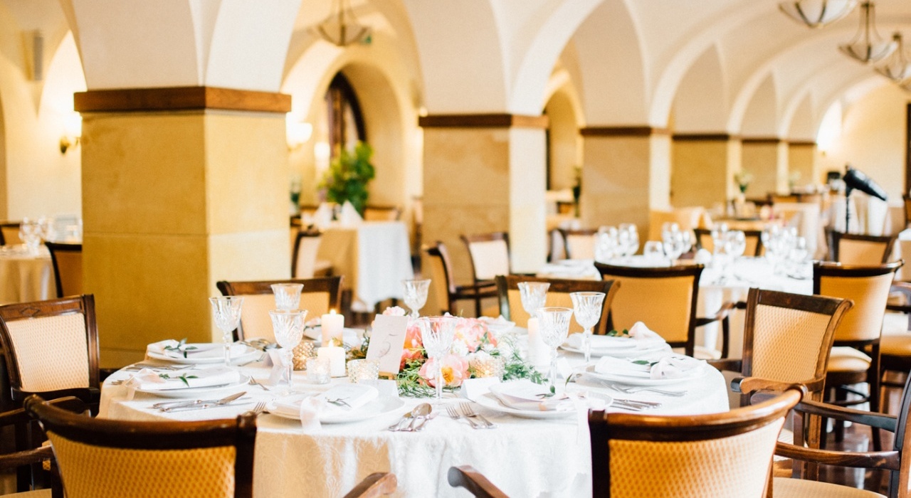 Elegantní jídelní sál s klenutými stropy a oblouky, lustry, kulatý stůl prostřený křišťálovými sklenicemi, svíčkami a květinovou dekorací.