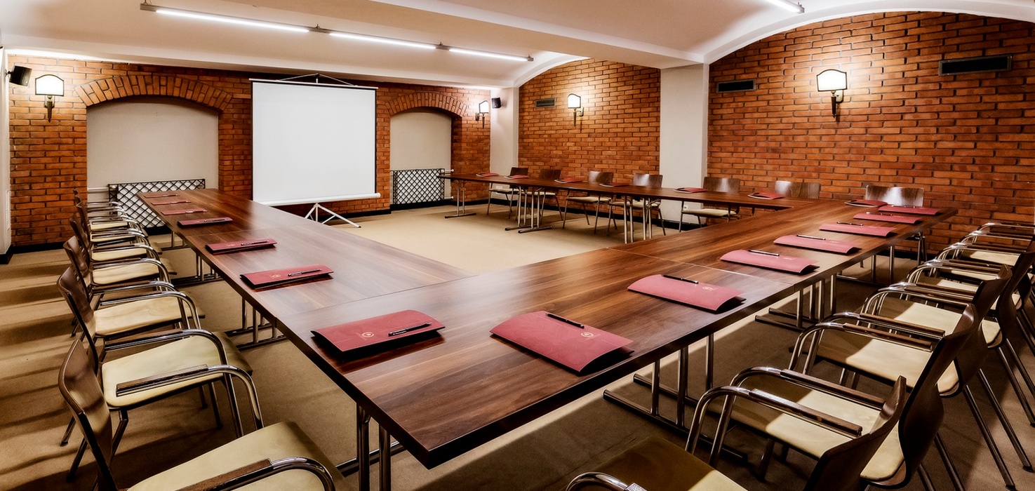 U-shaped hotel meeting room with wooden tables, padded chairs, projector screen, exposed brick walls and red folders with pens.