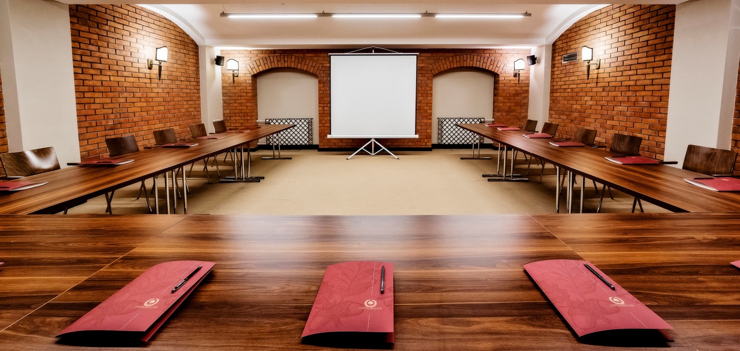 Conference room with U-shaped wooden tables, exposed brick walls, warm wall lamps, projector screen and red folders with pens.