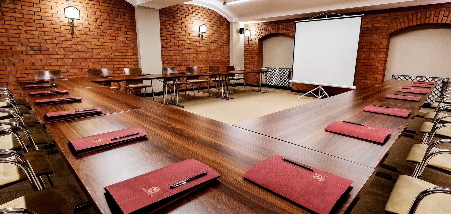 U-shaped conference room with wooden tables, brick walls, projection screen, chairs and red meeting folders with pens.