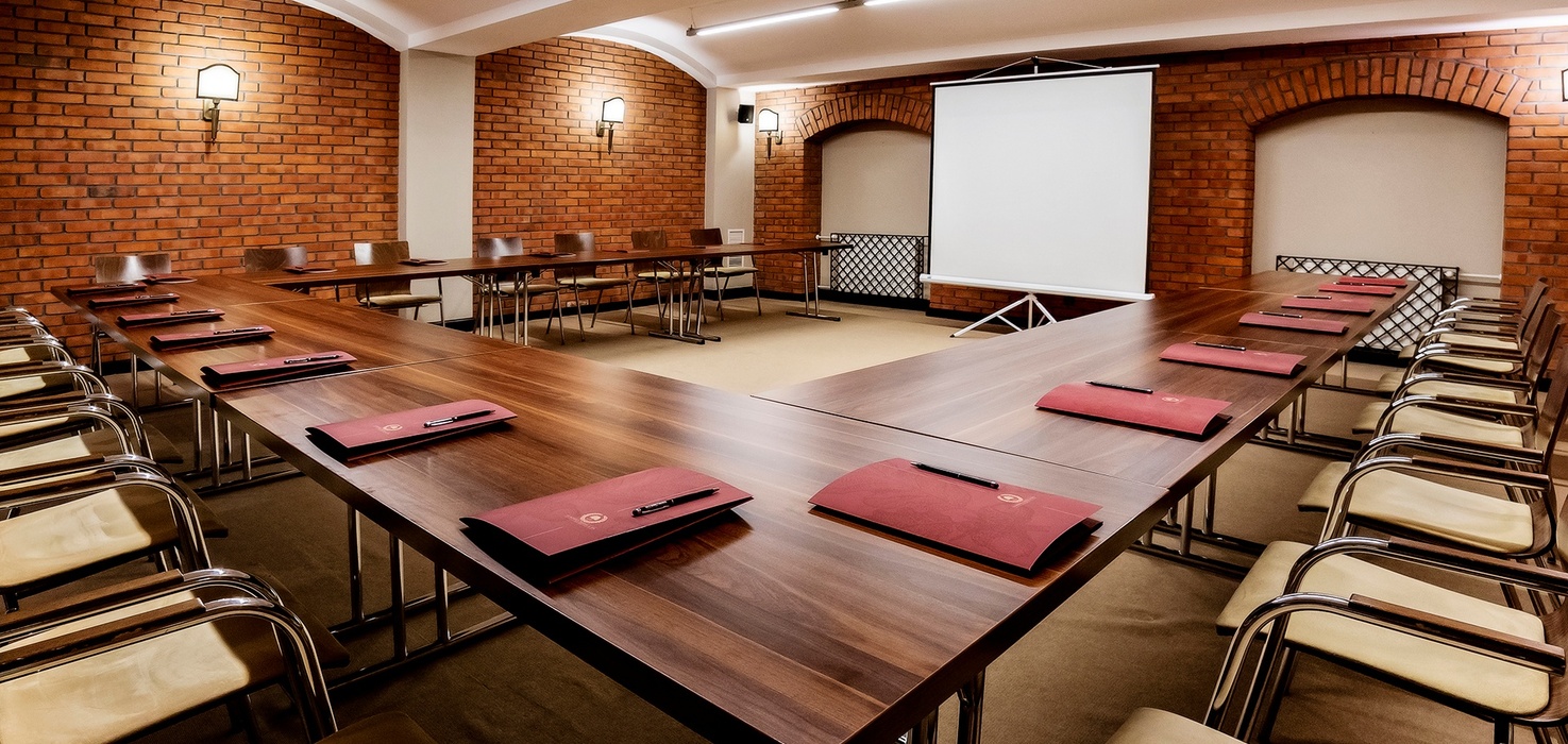 U-shaped meeting room with wooden tables, leather chairs, red notepads, projector screen and warm brick walls