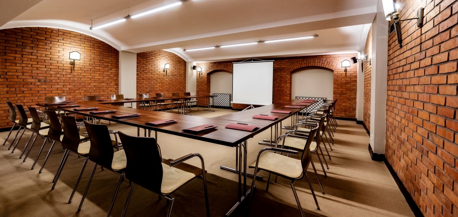 Brick-walled meeting room with U-shaped tables, chairs, projector screen and warm wall lighting