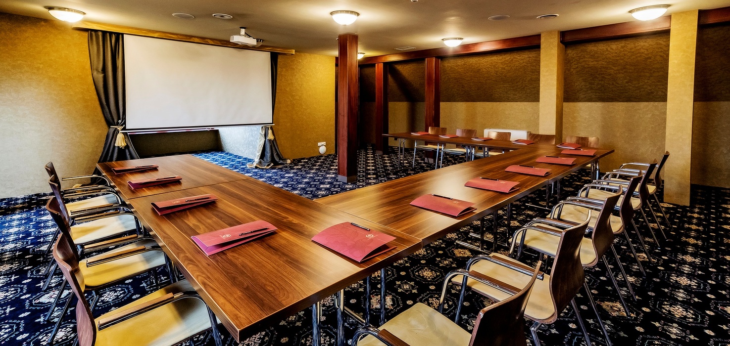 U-shaped hotel meeting room with wooden tables, padded chairs, projector and screen, note folders and warm lighting