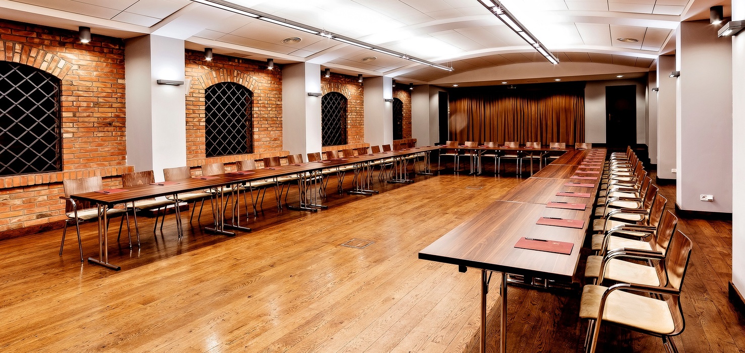 Spacious hotel conference room with U-shaped wooden tables, rows of chairs, exposed brick arched windows and warm ambient lighting
