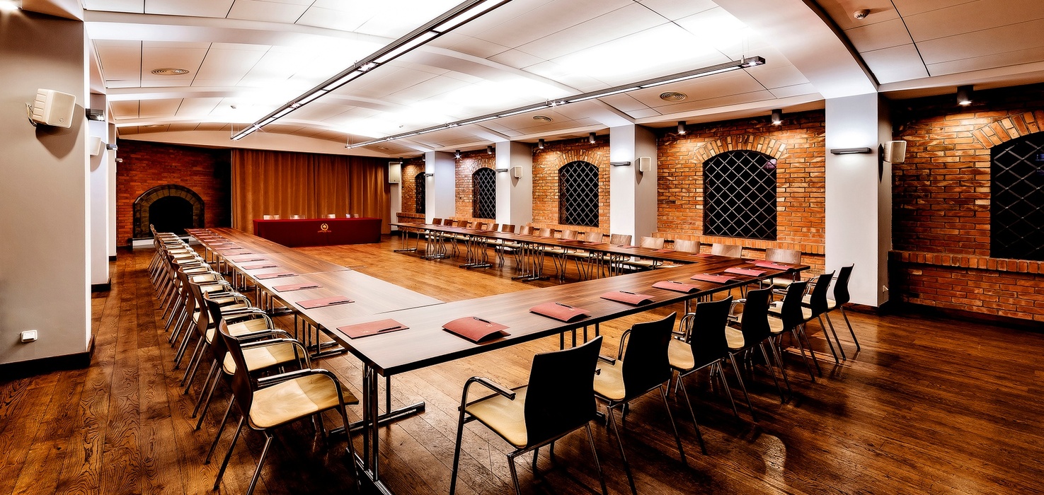 U-shaped conference room with warm wooden floors, exposed brick arched windows, long tables with notepads, ceiling lights, wall speakers and red-draped speaker table.