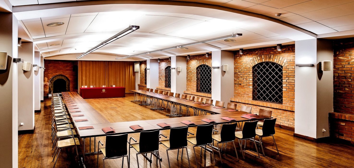 Spacious U-shaped conference room with wooden floors, exposed brick arched windows, long tables and stage