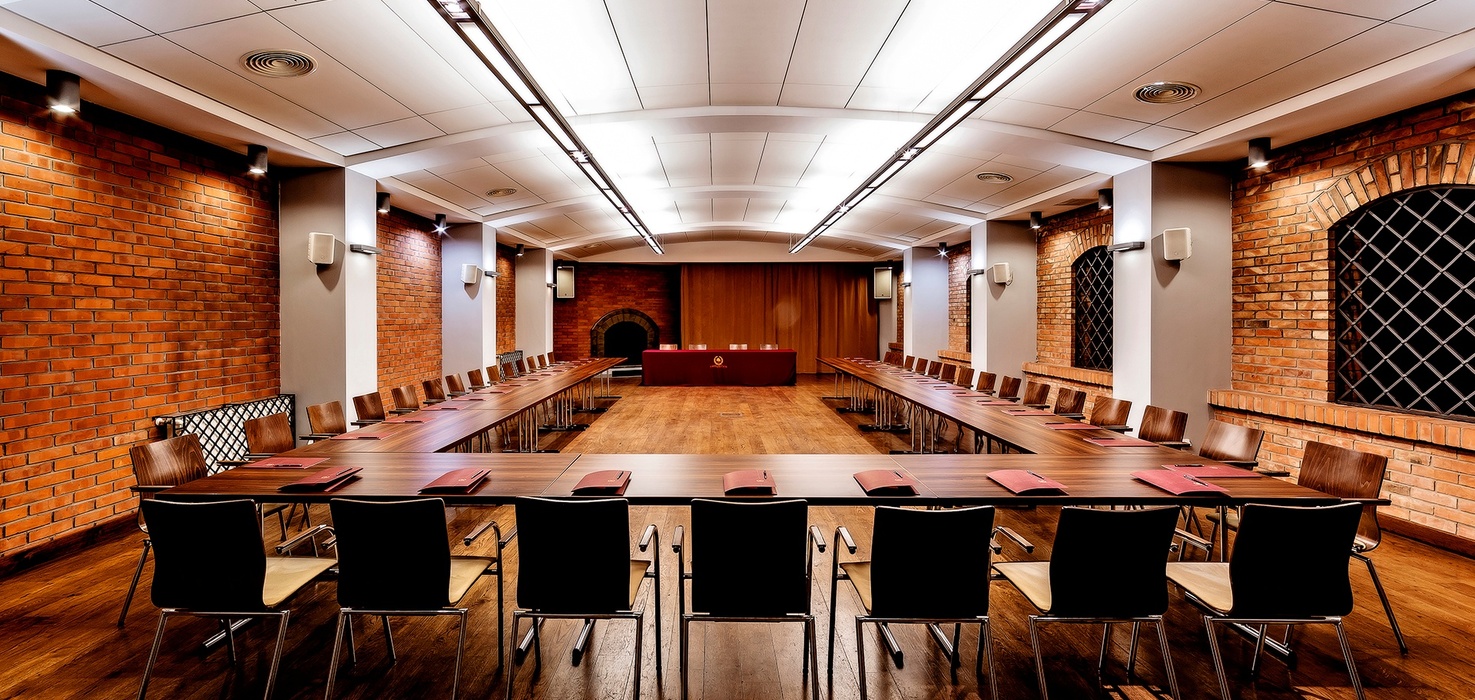Brick-walled meeting room with U-shaped wooden tables, arched grille windows, chairs, wood floor and front presentation table