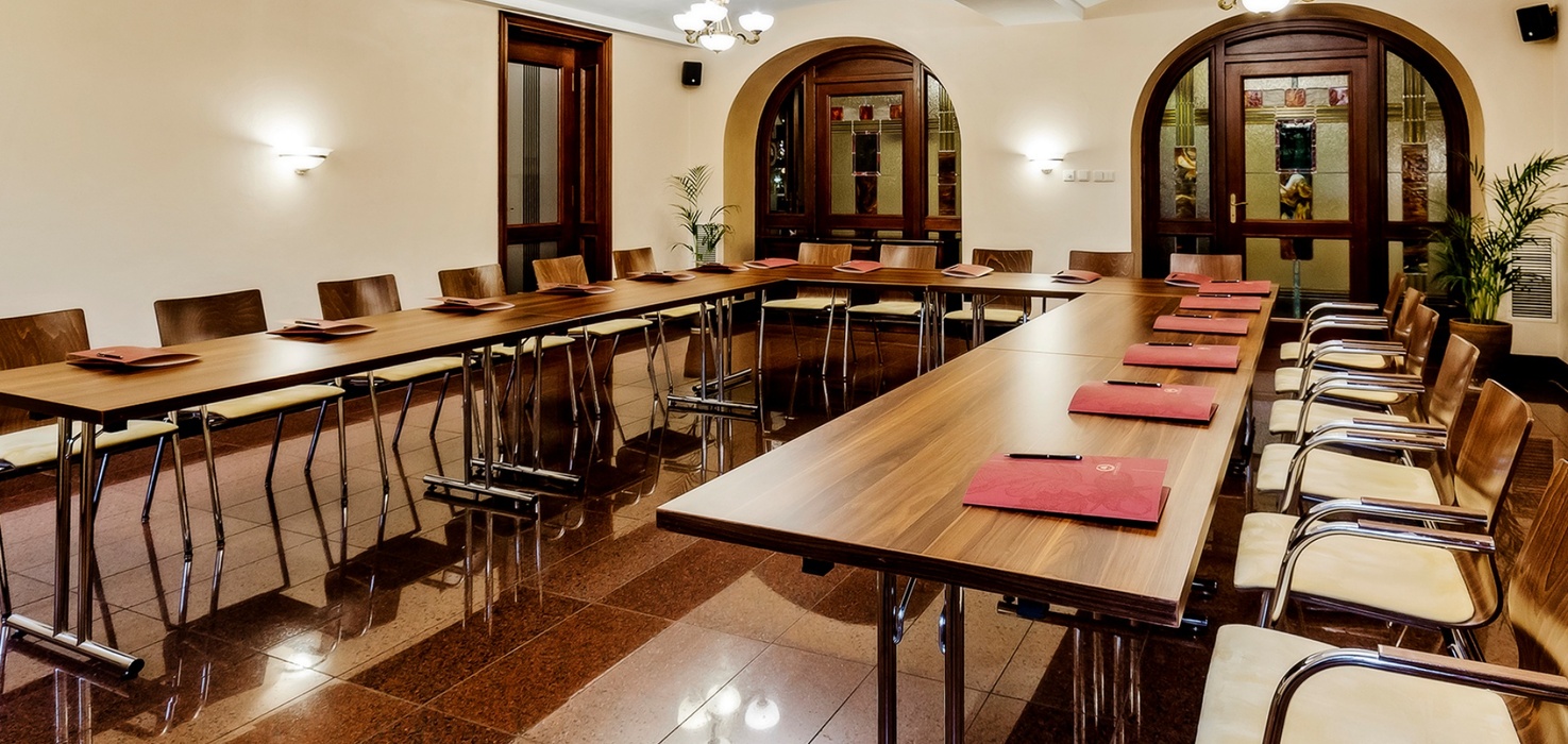 Konferenční místnost do U s dřevěnými stoly, koženými židlemi, zápisníky, obloukovými dveřmi s vitráží a lesklou kamennou podlahou
