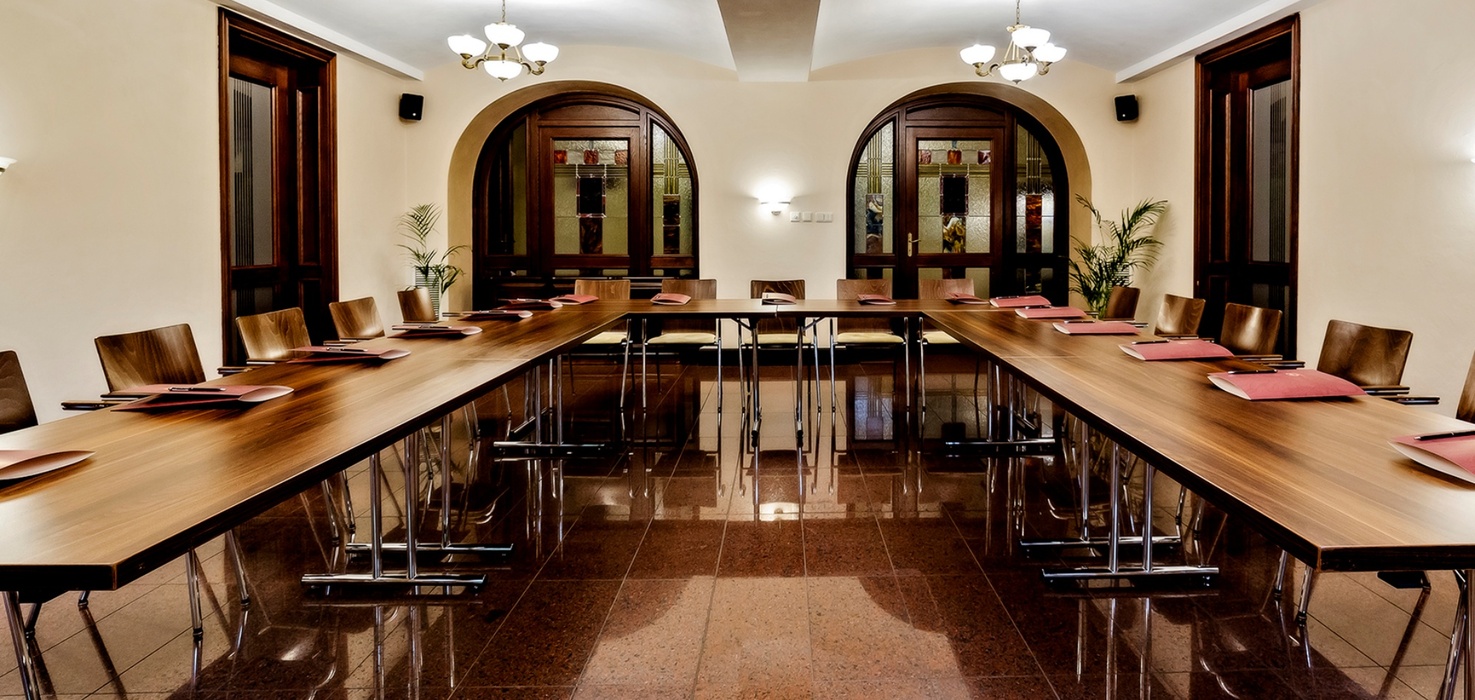 Elegantní hotelová zasedací místnost s U-tvarovanými dřevěnými stoly, koženými židlemi, lustry a vitrážními obloukovými dveřmi