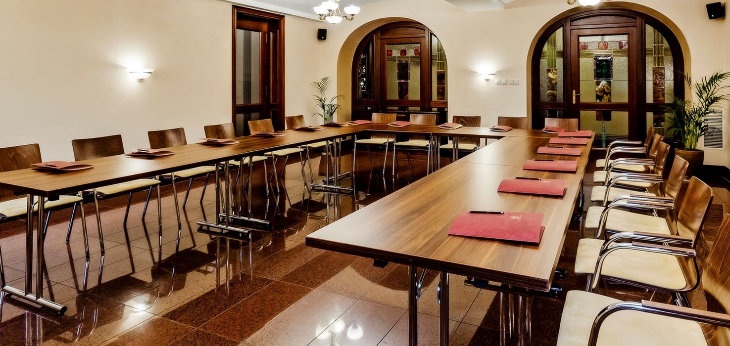 Konferenční místnost ve tvaru U s dřevěnými stoly, čalouněnými křesly, červenými složkami, lustry a obloukovými vitrážovými dveřmi