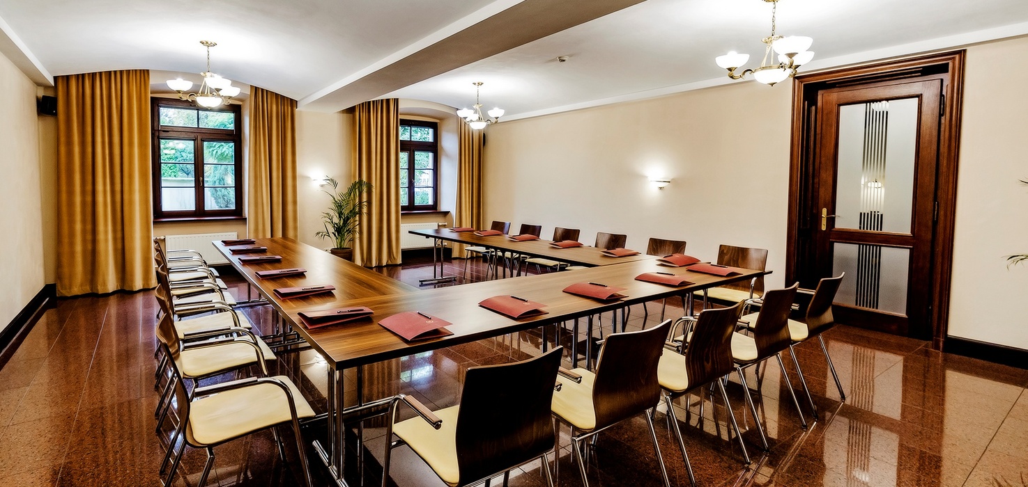 Konferenční místnost ve tvaru U s dřevěnými stoly a židlemi, červenými pořadači a pery, lustry, vysokými závěsy, okny a lesklou kamennou podlahou