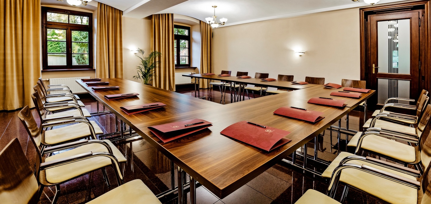Konferenční sál v hotelu ve tvaru U s dřevěnými stoly, čalouněnými židlemi, složkami a teplým osvětlením
