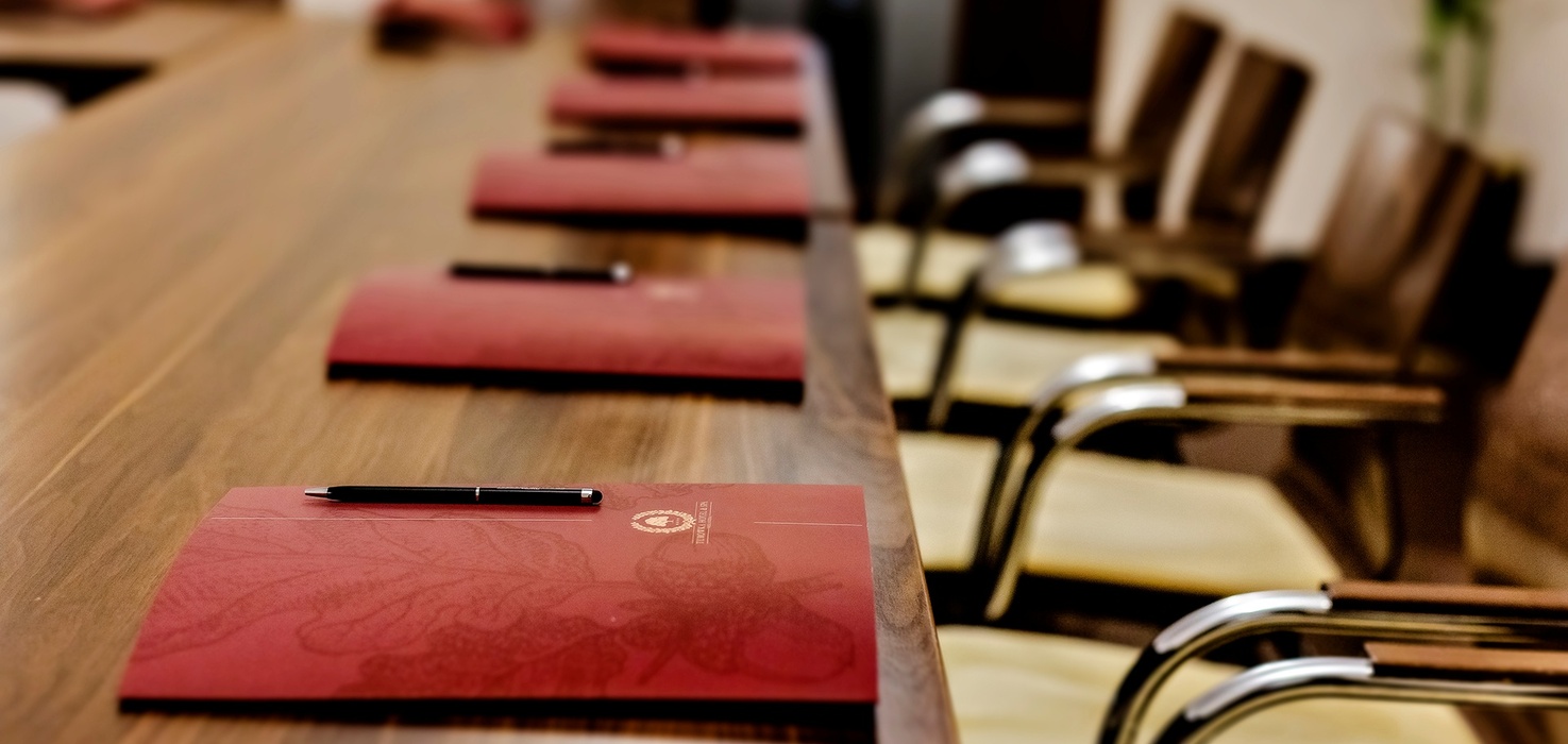 Dlouhý dřevěný konferenční stůl s červenými složkami a pery, pohodlná křesla připravená pro jednání