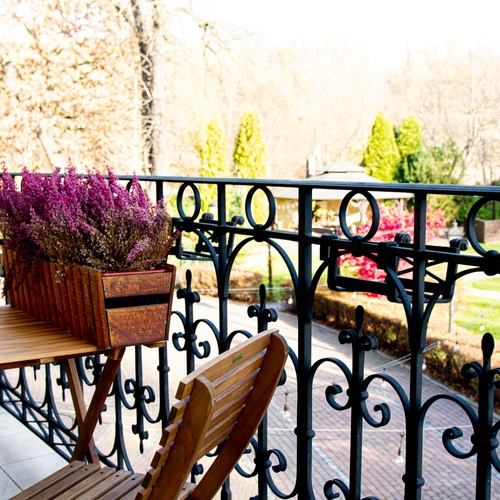 Kované zábradlí balkonu s dřevěným bistro stolem a židlemi, truhlík s fialovým vřesem, výhled na upravený park a altán.