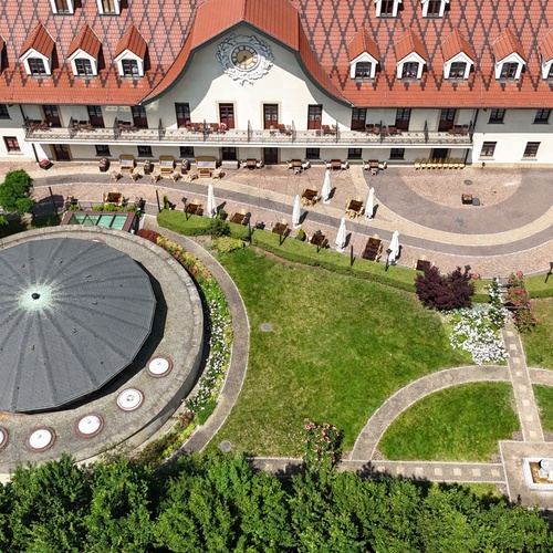 Historický hotel s červenou taškovou střechou a hodinami na fasádě, balkony a stolky na terase s výhledem na upravené kruhové zahrady a pavilon