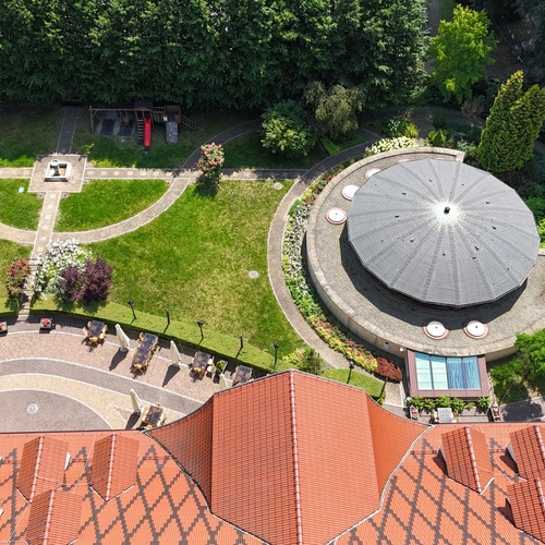 Letecký pohled na hotelové nádvoří s červenou taškovou střechou, kruhovým pavilonem, terasou s parasoly, upravenými trávníky, fontánou, altánkem a dětským hřištěm