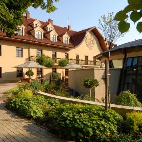 Malebné hotelové nádvoří se žlutou fasádou, červenou taškovou střechou, terasou s deštníky, posezením, upravenou zahradou a dlážděnou cestou.