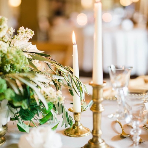 Lit tapered candles in brass holders beside greenery and glassware on an elegant wedding reception table.