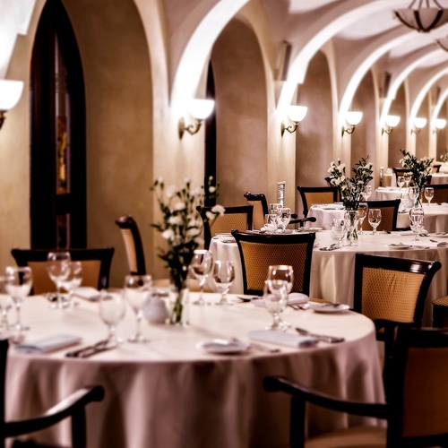 Elegant vaulted dining room with arched ceilings, warm wall sconces, round white‑clothed tables, floral centerpieces and fine glassware for intimate dining