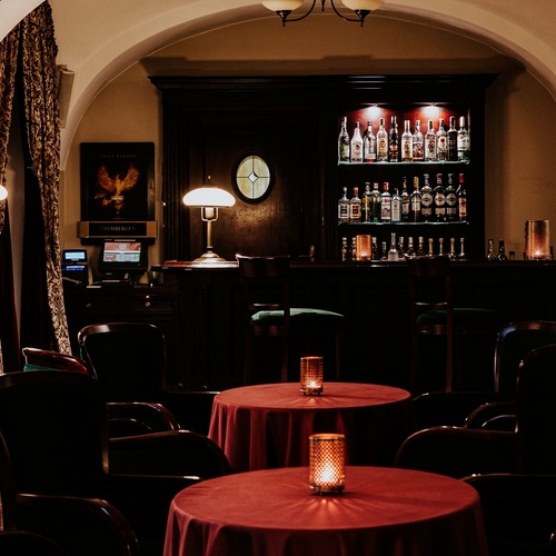 Útulný hotelový bar s obloukovým stropem, tmavým dřevěným barem, osvětlenými regály s alkoholem a kulatými stolky s červenými ubrusy.
