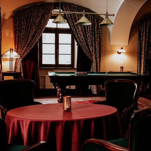 Elegantní hotelový salonek s kulečníkem, klenutými stropy, vzorovanými závěsy, teplým osvětlením a kulatým stolem s bordó ubrusem.
