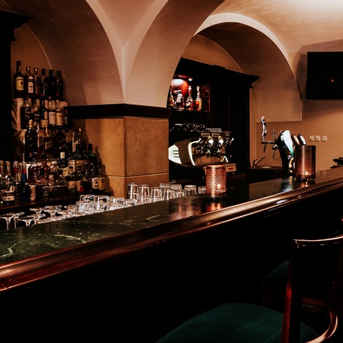 Příjemný hotelový bar s klenutými stropy, tmavým dřevěným pultem, podsvícenými lahvemi, výčepem, sklenicemi a tlumeným osvětlením.