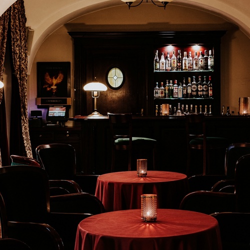 Cozy hotel lounge with arched ceiling, dark wood bar with illuminated bottles, round tables with red cloths and candle lanterns