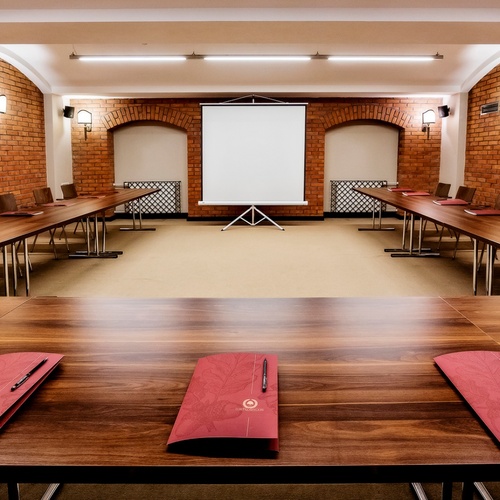Konferenční místnost v uspořádání do U s dřevěnými stoly, cihlovými stěnami, teplým osvětlením, projekčním plátnem a červenými složkami s pery.