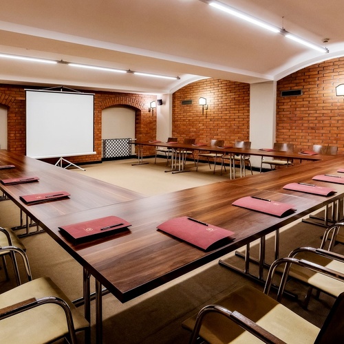 Konferenční místnost ve tvaru U s dřevěnými stoly, koženými židlemi, projekční plachtou a teplými cihlovými zdmi