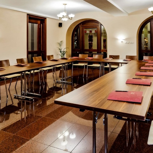 Konferenční místnost ve tvaru U s dřevěnými stoly, čalouněnými křesly, červenými složkami, lustry a obloukovými vitrážovými dveřmi
