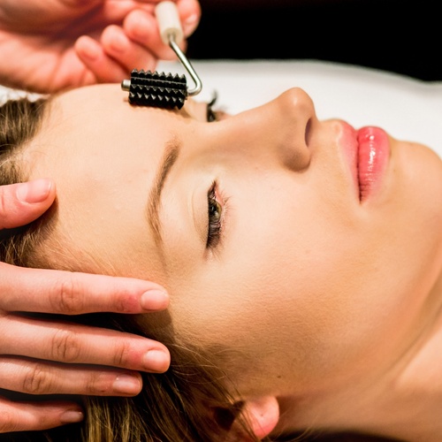 Žena při relaxačním spa ošetření obličeje s vroubkovaným válečkem na čele, ruce terapeuta jemně podepírají hlavu.
