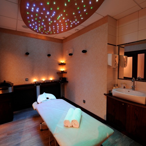 Relaxační spa místnost s masážním stolem, srolovanými ručníky, umyvadlem, svíčkami a hvězdným LED stropem.