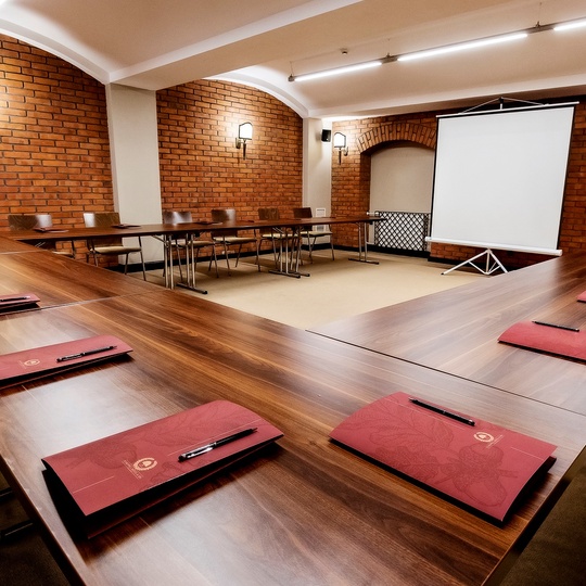 Konferenzraum in U‑Form mit Holztischen, roten Ordnern und Stiften, sichtbaren Ziegelbögen, Projektionsleinwand und warmer Wandbeleuchtung
