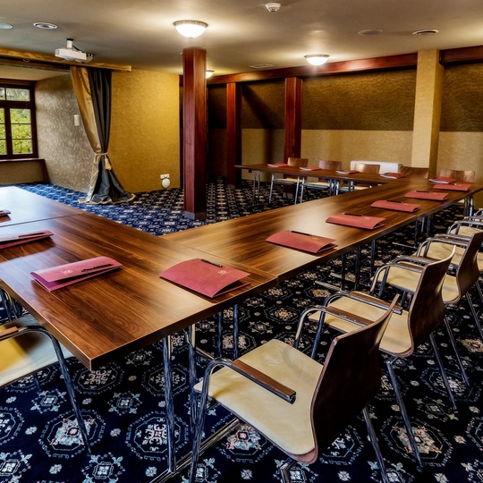 Konferenzraum in U-Form mit Holztischen, gepolsterten Stühlen, roten Ordnern an jedem Platz, Deckenprojektor und Fensterblick ins Grün.