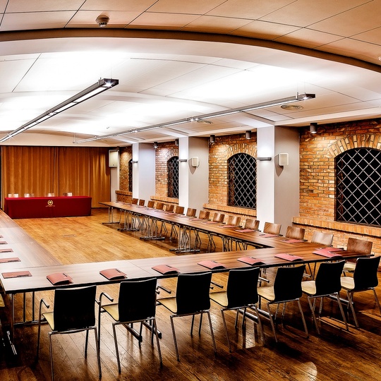 Geräumiger Konferenzraum in U-Form, Holzboden, freiliegende Backsteinbögen mit Gitterfenstern, lange Tische und Bühne