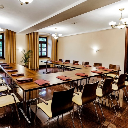 Konferenzraum in U-Form mit Holztischen, cremegepolsterten Stühlen, roten Unterlagen, Kronleuchtern, großen Fenstern mit goldenen Vorhängen und glänzendem Boden