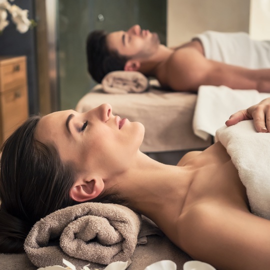 Couple enjoying side-by-side spa massages on padded beds, with candles and rolled towels for a calm atmosphere.