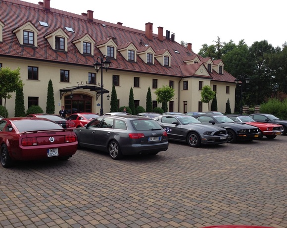 Hotel façade with red-tiled roof, canopy entrance, manicured shrubs and cobblestone forecourt lined with parked sports cars