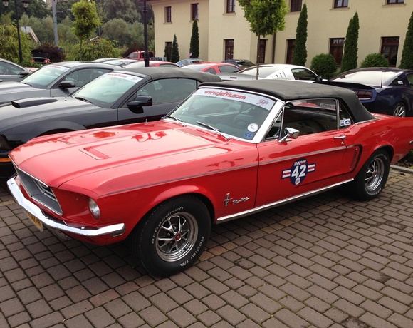 Red 1967 Ford Mustang convertible with racing decal parked on hotel's cobblestone forecourt, hotel façade and trees in the background